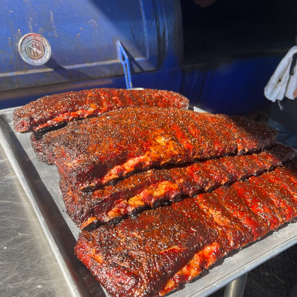 A tray of perfectly smoked, seasoned ribs with a rich, dark crust, next to a blue smoker.