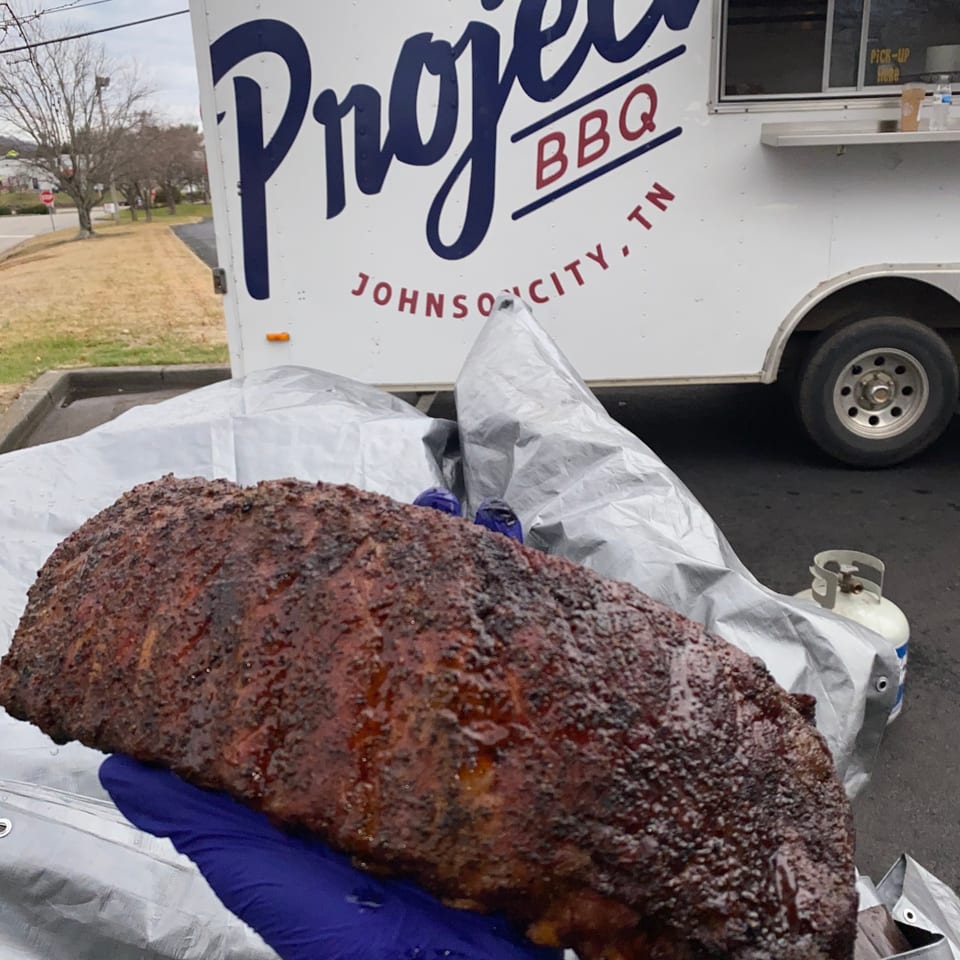 A hand in a blue glove holds a rack of Project BBQ ribs in front of their food truck.