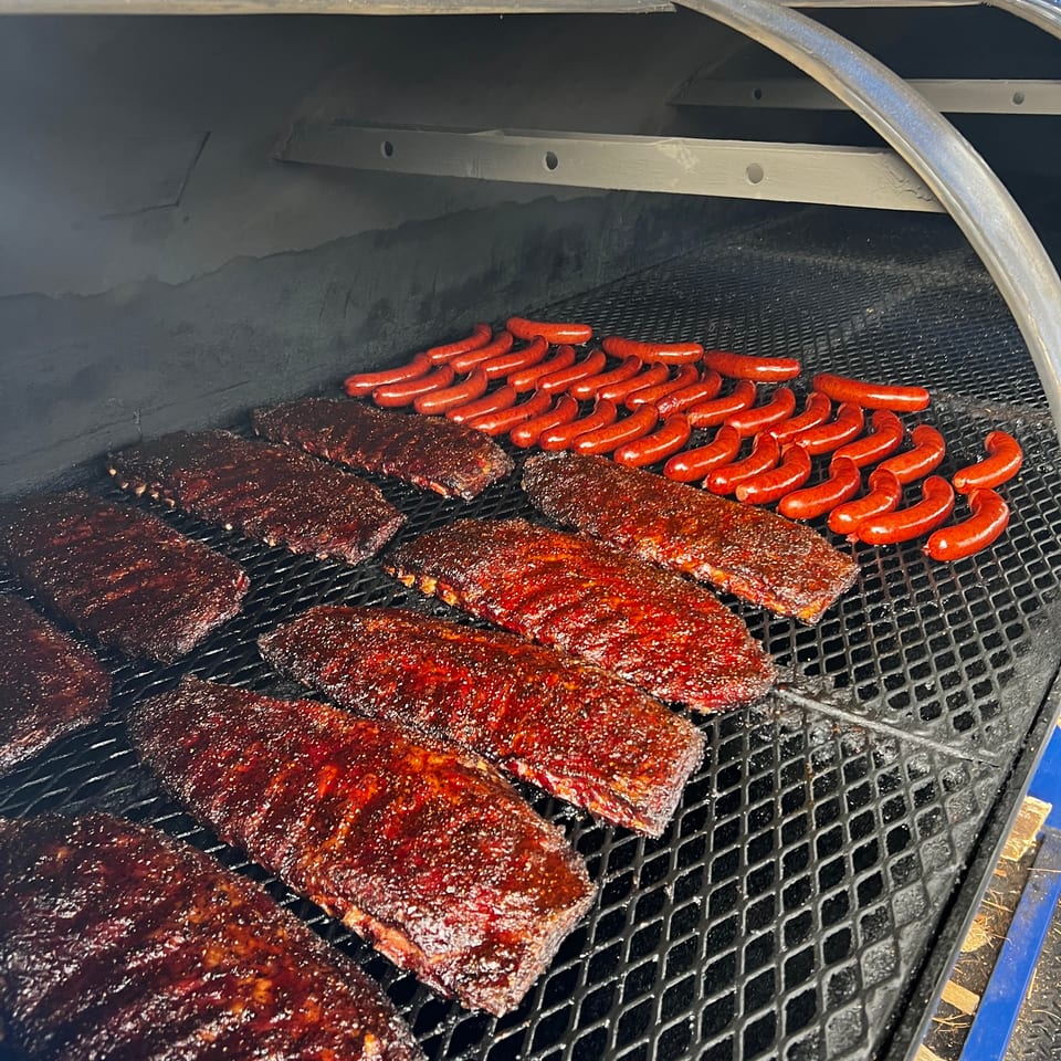 Rows of glistening BBQ ribs and bright red sausages cooking on a large grill.