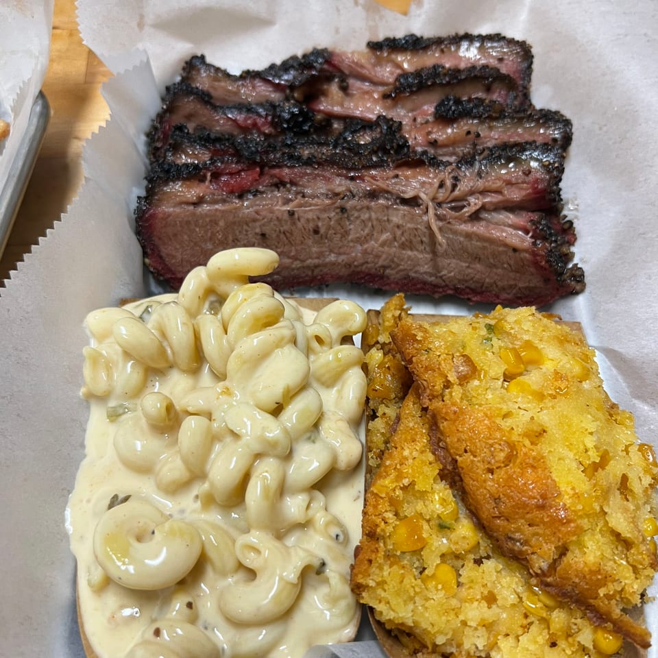 Sliced brisket with mac and cheese and cornbread in a paper-lined tray.