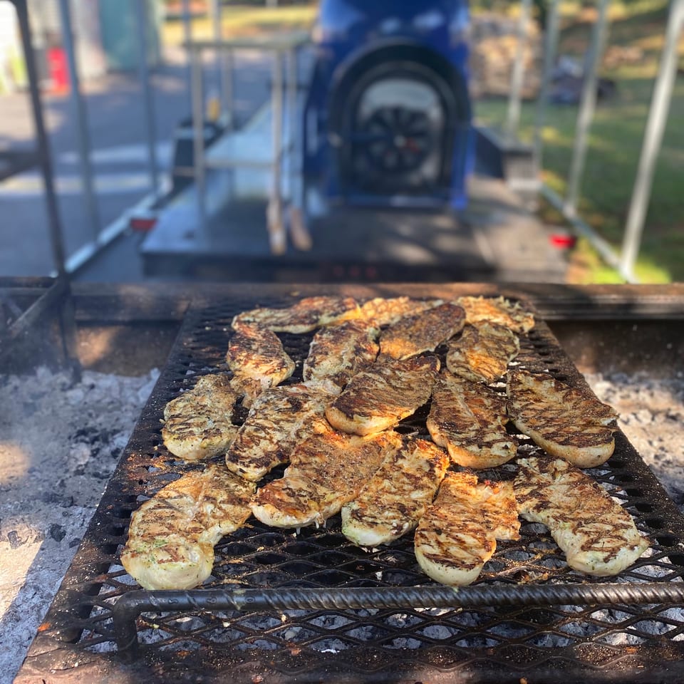 Grilled chicken breasts cooking on a large outdoor barbecue grill with a blue smoker in the background.