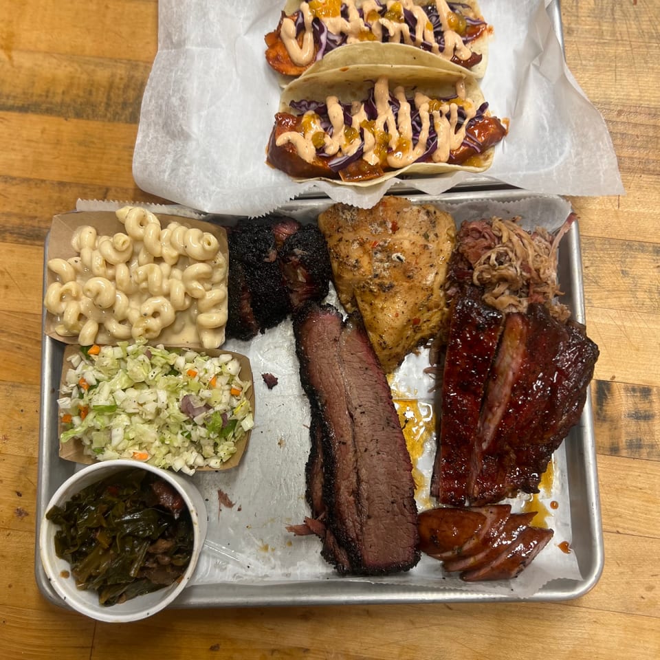 A metal tray with various BBQ meats, sides, and two tacos on a wooden table.